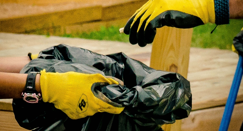 Gloved hands hold a trash bag while another gloved hand drops something inside. 